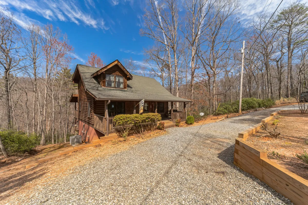 This image displays the wooden cabin surrounded by tall trees, with a gravel driveway leading up to it. The lush natural setting and visible outdoor space evoke a sense of peaceful seclusion and direct access to nature.