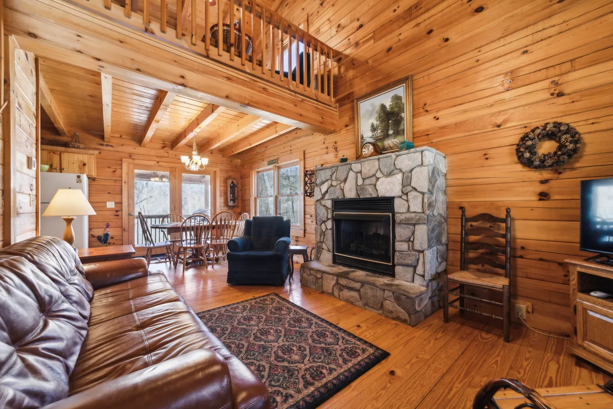 This image captures the heart of the Great Room, featuring a commanding stone fireplace, a plush leather sofa, a spacious wooden dining table, and a cozy blue armchair atop a patterned rug. The arrangement is perfect for family gatherings, playing games on the table, or relaxing by the fire.