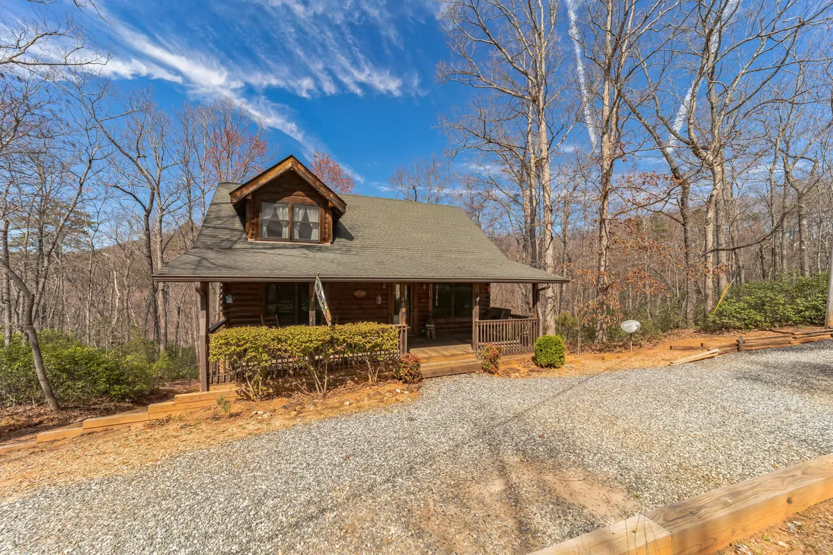 The facade of a log cabin features a welcoming front porch and a gravel driveway, framed by tall trees. The scene feels cozy and secure, emphasizing both the inviting outdoor space and the secluded, wooded environment.