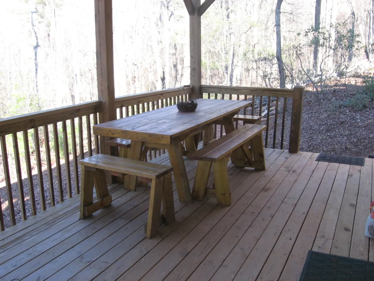 A spacious wooden table with benches is set on an outdoor patio, surrounded by greenery and trees. The scene feels communal, tranquil, and ideal for shared moments in nature.