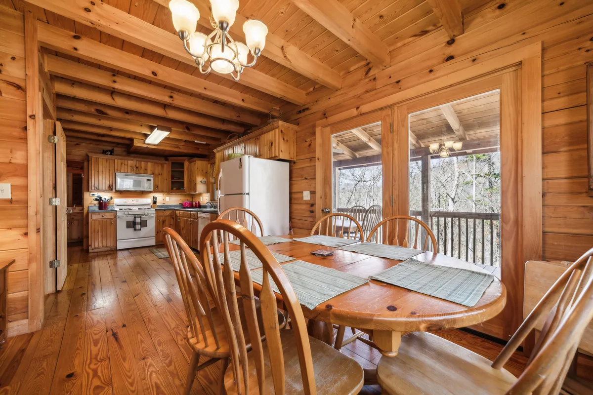 A bright, inviting dining area with a substantial wooden table, matching chairs, and visible kitchen appliances in the background. Large windows allow natural light to flood the space, creating the perfect setting for shared meals, card games, or simply enjoying the view while eating.