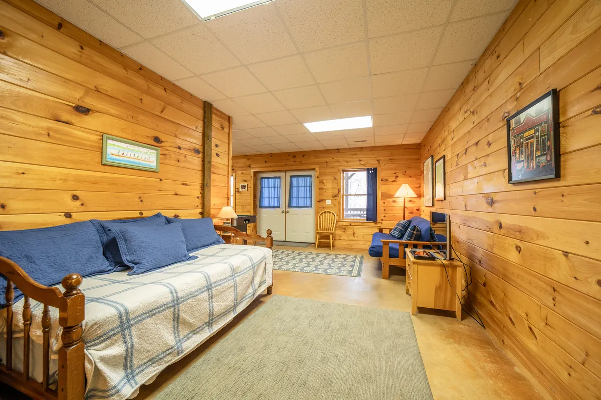 A warm, open living area with wood-paneled walls, a sofa bed with blue cushions, a softly lit lamp, dining chair, and a patterned rug. The layout is inviting and hints at the flexibility for additional guests, reflecting the loft or basement sleeping arrangements.