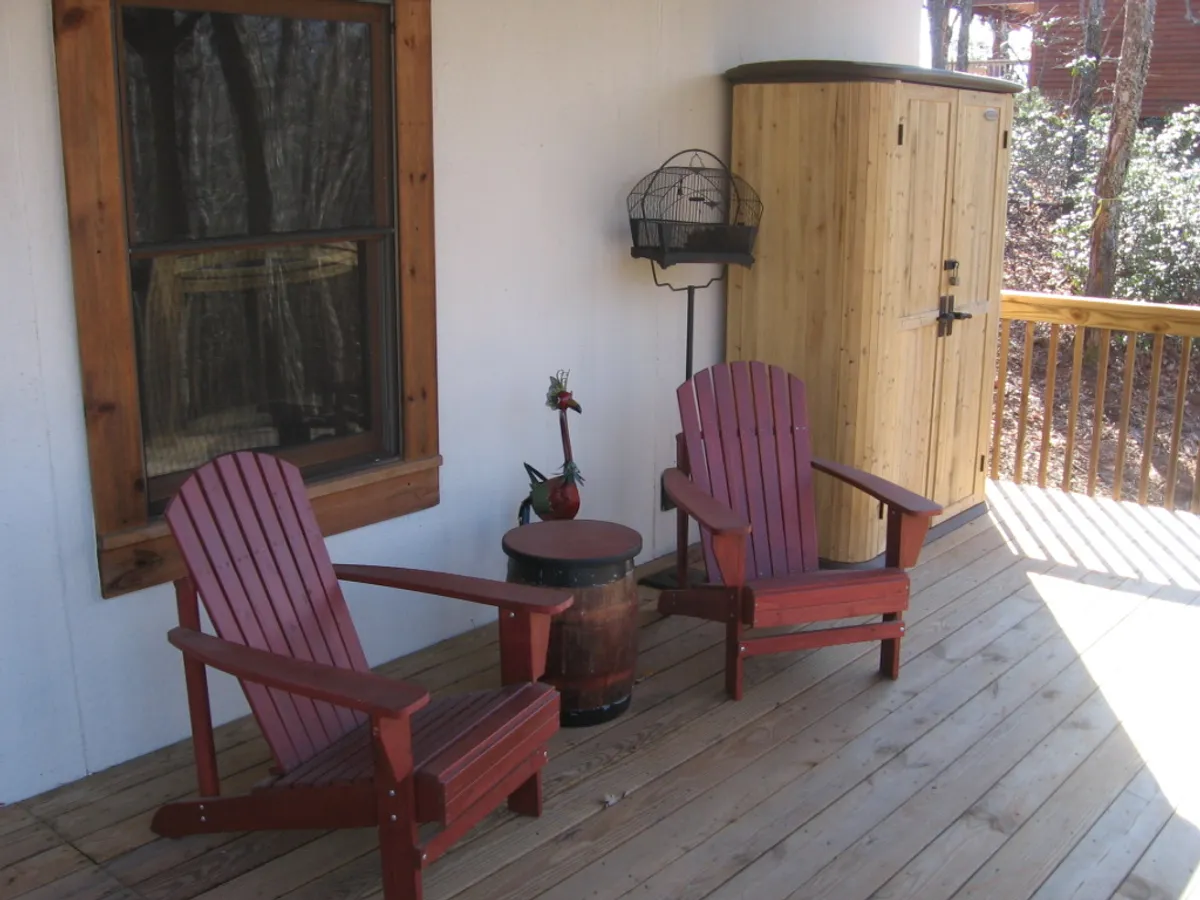 The image features red wooden chairs and a barrel table set near a wooden storage unit and a birdcage. The arrangement suggests a cozy outdoor or transitional indoor-outdoor space, ideal for relaxing, chatting, or enjoying nature. The soft lighting and rustic decor evoke a tranquil, inviting atmosphere that feels connected to the outdoors.