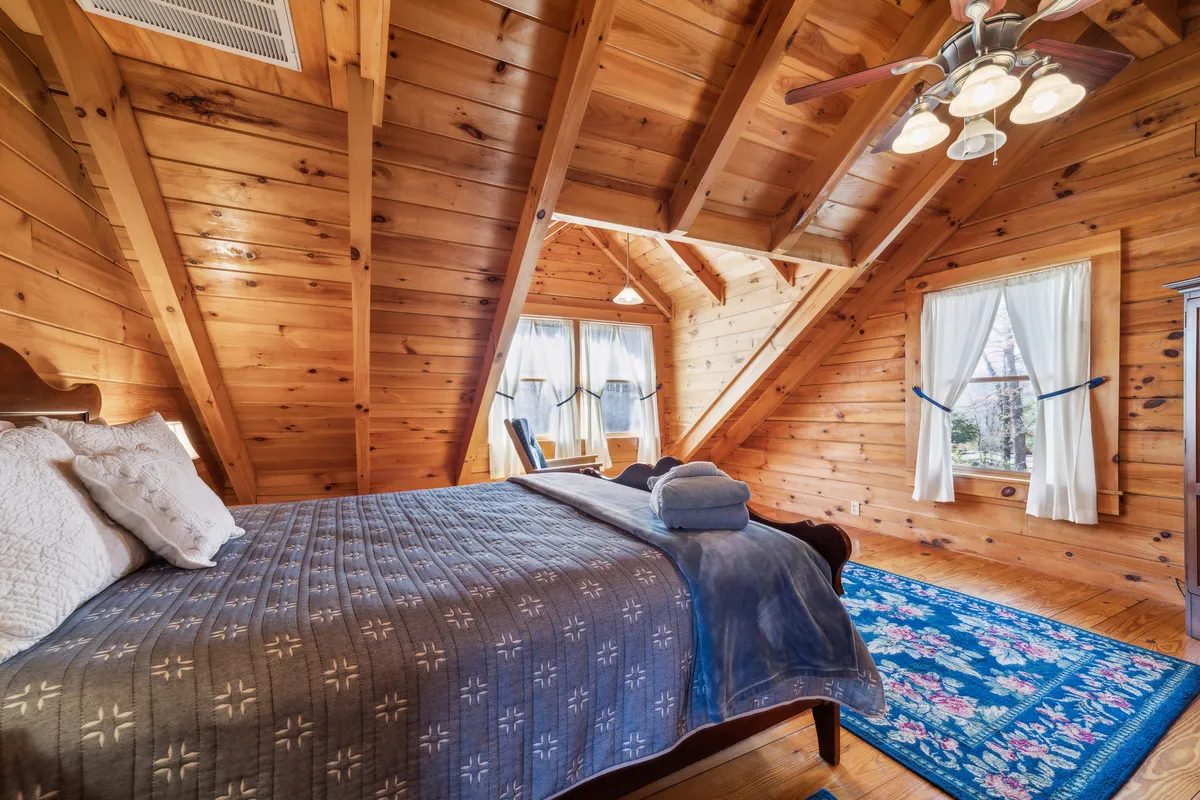 This image showcases a tranquil loft or bedroom with exposed wooden beams, a cozy plaid bedspread, a blue rug, and large windows that let in ample natural light. The inviting setup suggests a peaceful retreat, ideal for reading, napping, and enjoying serene moments together.
