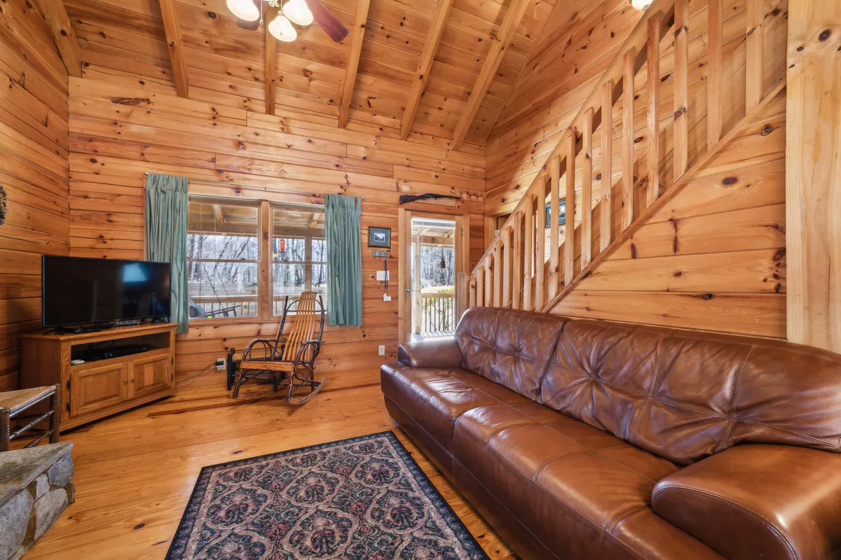 A warm, welcoming living area with a leather sofa, wooden staircase, TV unit, and rocking chair. Large windows flood the space with natural light, offering views of the surrounding scenery. The environment is spacious and relaxed, suitable for gathering, enjoying casual conversations, or simply soaking in the view.