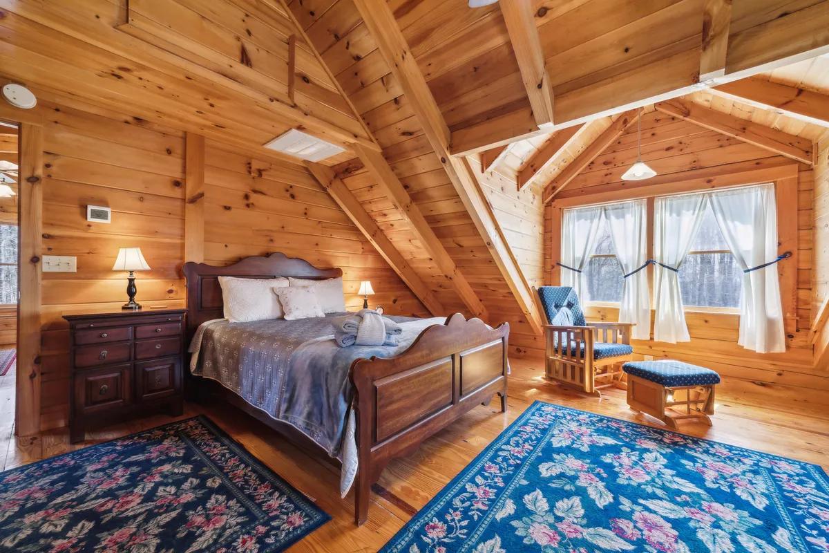 A tranquil master bedroom with a wooden bed frame, intricate headboard, soft area rug, and large windows draped with sheer curtains. The scene is serene, accented by natural light and warm textures, suggesting a peaceful retreat.