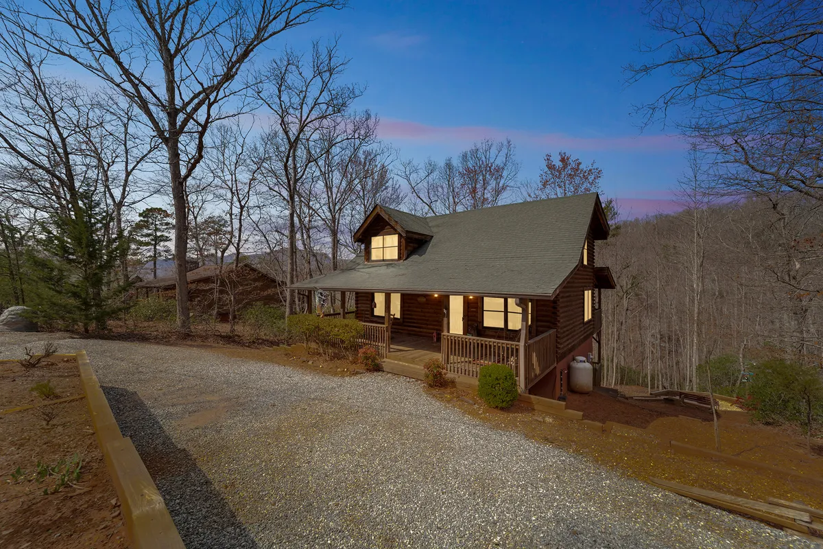 The hero image showcases a classic log cabin nestled among towering trees, with a welcoming covered front porch adorned with rocking chairs. The gravel driveway leads up to the cabin, suggesting easy access and a sense of arrival. The surrounding woods create an atmosphere of privacy and tranquility, hinting at opportunities for outdoor relaxation and nature experiences.