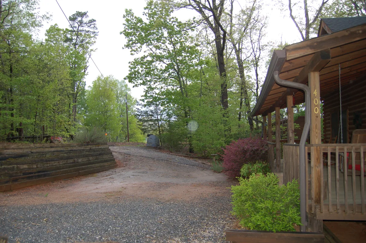 A peaceful front porch with rocking chairs looks out onto a gravel path, surrounded by mature trees and landscaped bushes. The covered porch is set against the backdrop of a private, wooded lot, evoking tranquility and a connection to nature.
